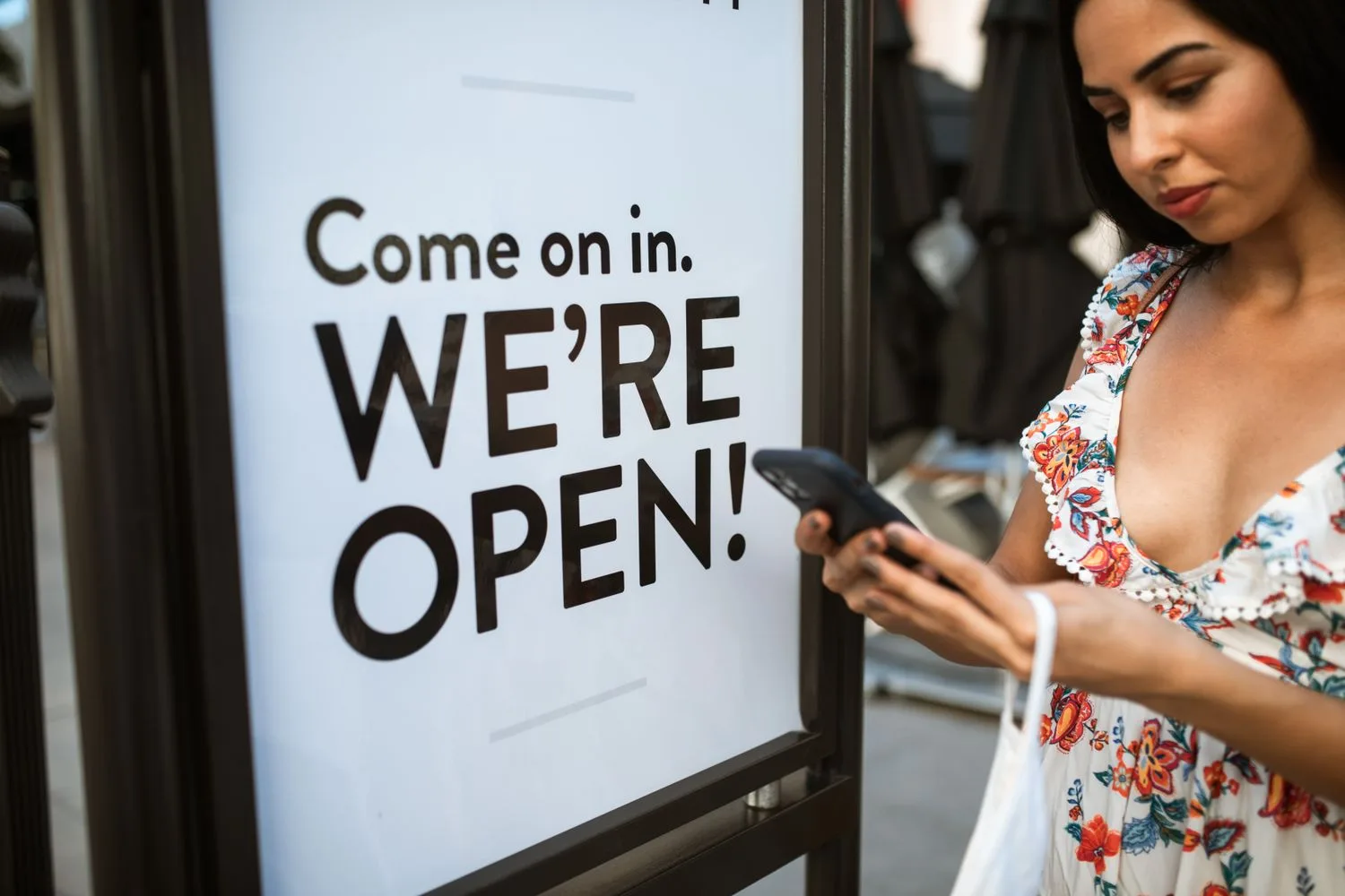 A woman looking at her phone beside a shop door sign that reads “Come on in. We're open!”