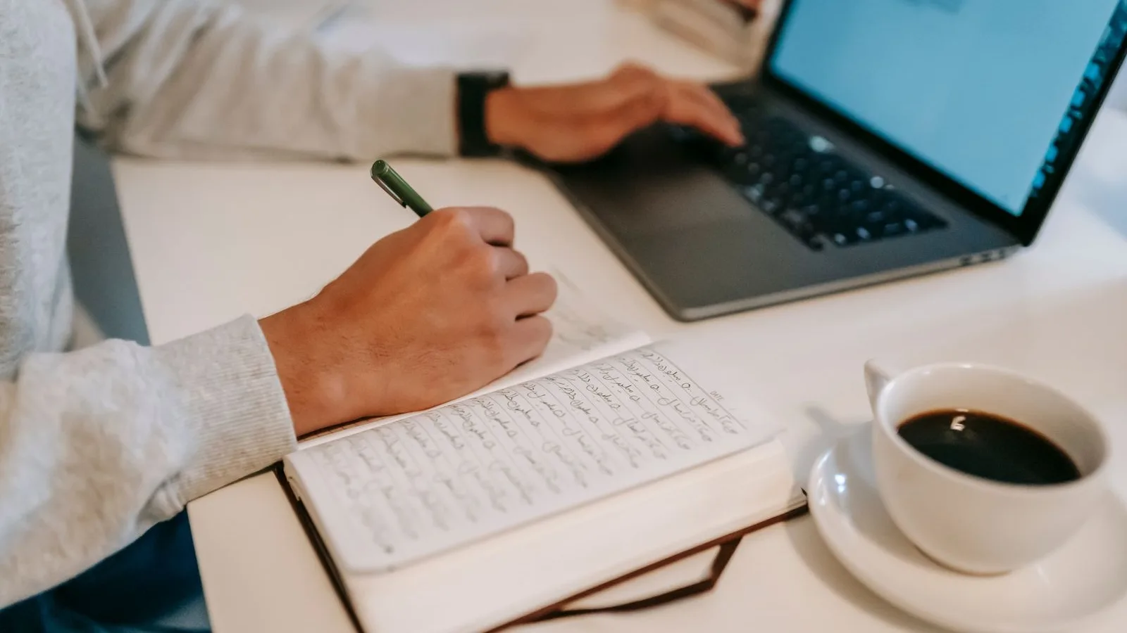 A hand taking notes with a pen in a notebook next to a laptop and a coffee, like someone checking a text by hand before publishing it