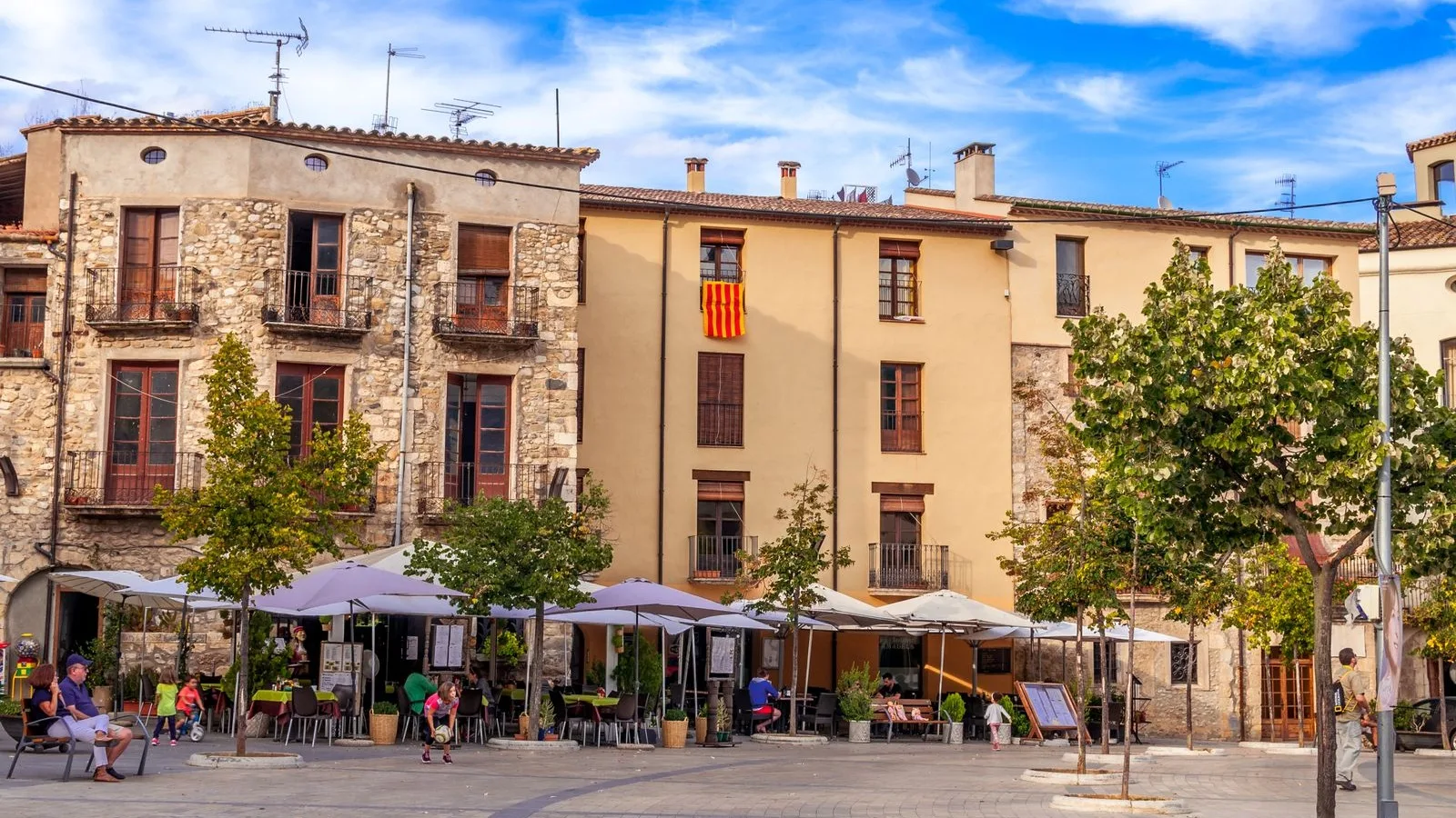 Plaça d'un poble català amb la senyera i terrasses de cafè plenes, el comerç local que viu de ser trobat