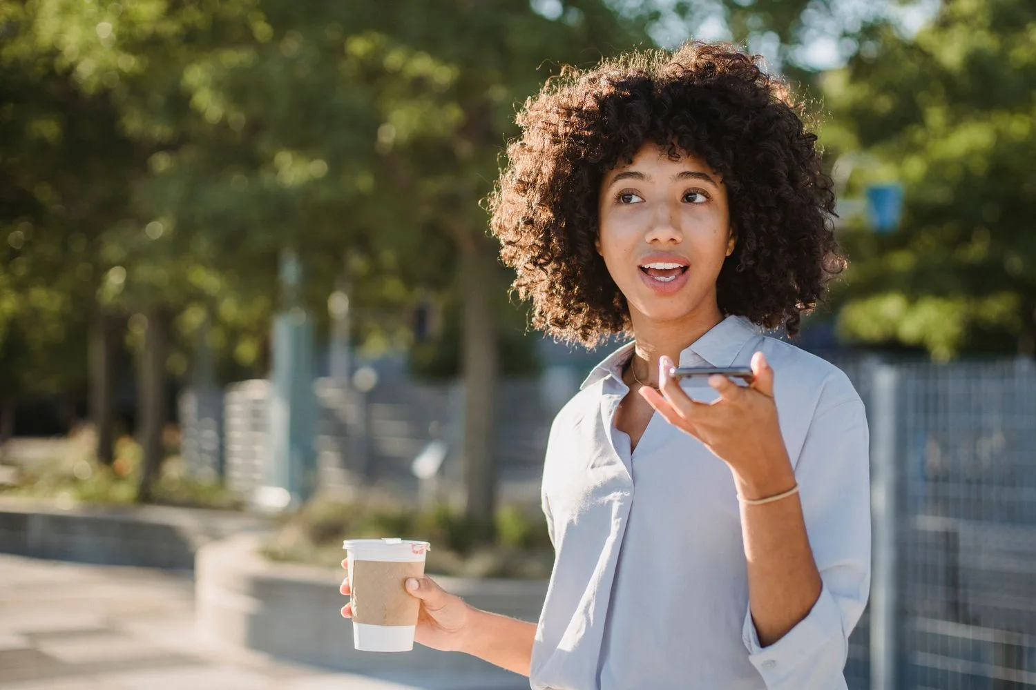 Una mujer habla al móvil por la calle con un café en la mano, como quien le pregunta algo a un asistente